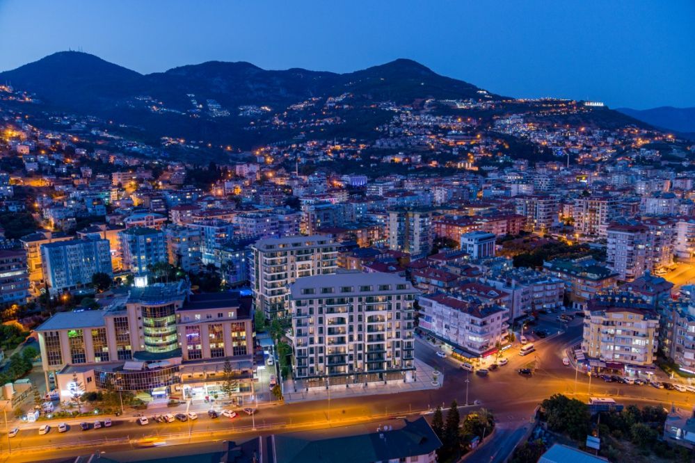 Akşam ışıklarıyla Alanya şehir manzarası sunan modern konut projesi, estetik mimarisiyle öne çıkıyor.Kleopatra Plajı’na Yürüme Mesafesinde Modern Konut Projesi